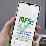 Mão segurando um smartphone que exibe a tela inicial do aplicativo do Módulo Emissor da nova Nota Fiscal de Serviço Eletrônica (NFS-e) Nacional, com um computador e máquina de cartão ao fundo.