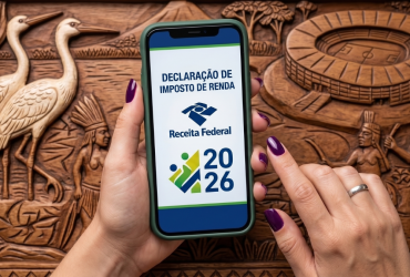IR 2026: só 45% dos cuiabanos que declararam no ano passado já entregaram; prazo acaba em 29 de maio Mãos segurando um smartphone que exibe o aplicativo da Receita Federal para a Declaração de Imposto de Renda 2026. Ao fundo, um entalhe em madeira com figuras indígenas, garças e um estádio de futebol.